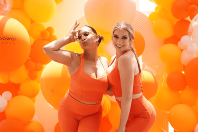 Two women in orange activewear pose and smile in front of a vibrant Los Angeles backdrop decorated with stunning orange and yellow balloon installations.