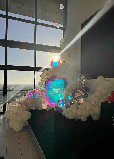 A stunning balloon installation of white and iridescent balloons is set up near large windows with sunlight streaming through, illuminating the scene in true Los Angeles style.