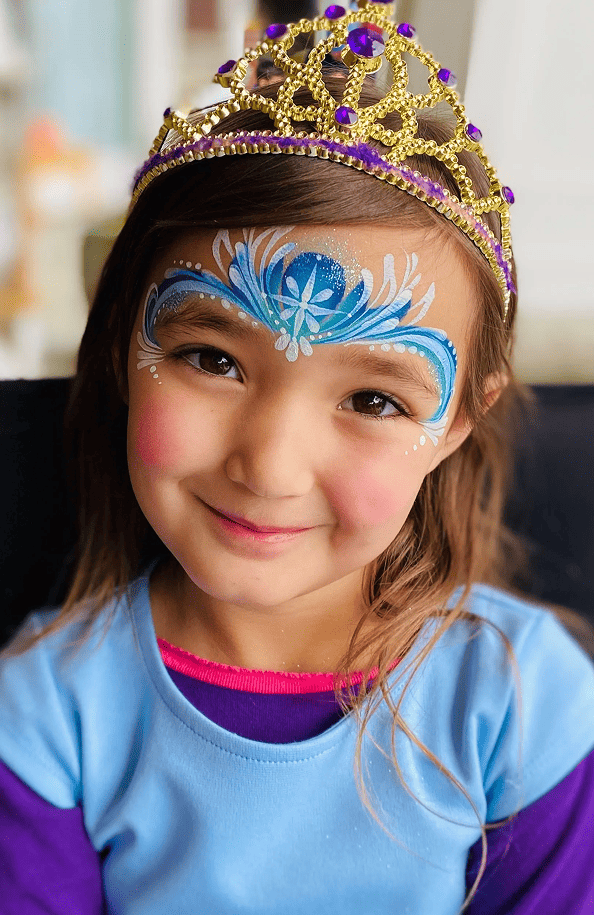 Young child wearing a gold and purple crown, blue and purple face paint, and a blue shirt, smiling at the camera at a Los Angeles event featuring creative balloon installations.