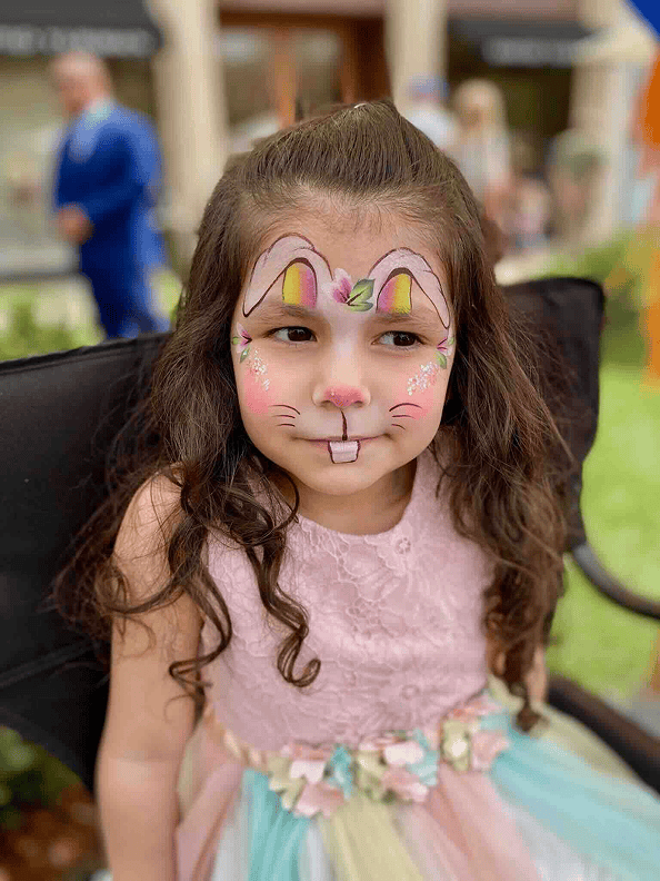 Young girl with long brown hair sits outdoors wearing a pastel dress, her face painted like a bunny with ears, whiskers, nose, and flowers on her cheeks, surrounded by colorful balloons from a talented balloon artist.