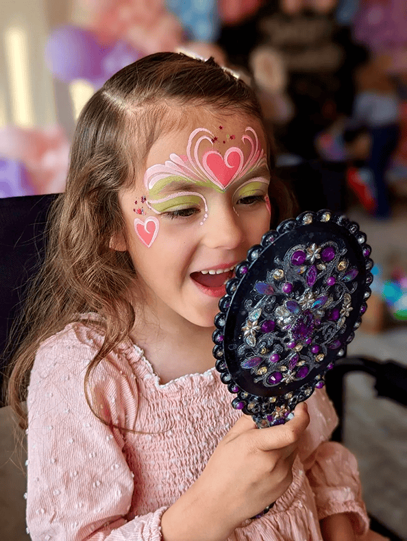 Young girl with decorative face paint featuring hearts and swirls smiles while looking into a jeweled hand mirror, as colorful balloon decor by a talented balloon artist surrounds her.