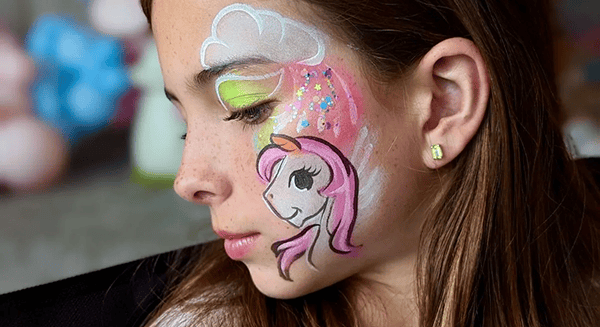 A child with unicorn and rainbow face paint, featuring pink, white, and green colors, looks downward with eyes closed at a festive corporate event adorned with vibrant balloon decor.