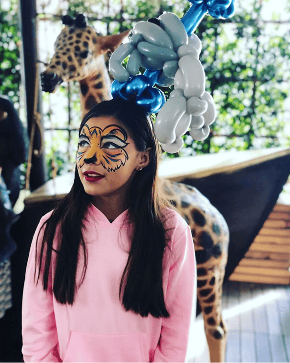 A girl in a pink hoodie with tiger face paint and a balloon hat, made by a talented balloon artist, stands in front of a giraffe statue in Los Angeles.