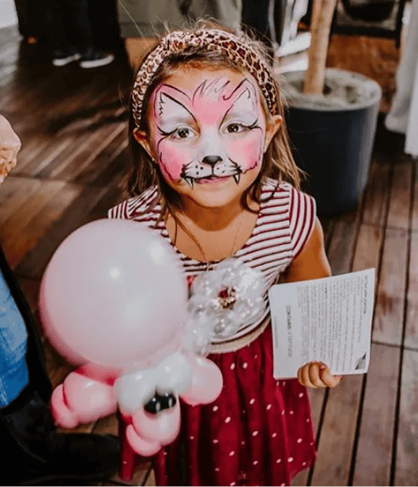 Young girl with fox face paint holds a pink balloon animal and a piece of paper, wearing a red dress with white stripes, indoors on a wooden floor surrounded by colorful balloon decor.