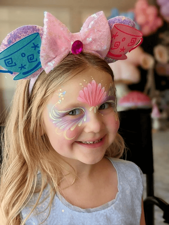 Smiling young girl with colorful face paint and large pink bow headband with blue and pink ears, wearing a light blue dress, holds playful balloons from a talented balloon artist.