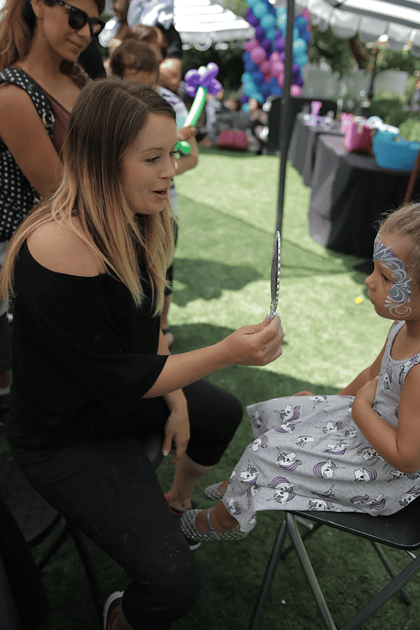 A woman holds a mirror for a young girl with face paint at an outdoor Los Angeles corporate event, while other people, decorations, and a balloon artist are visible in the background.
