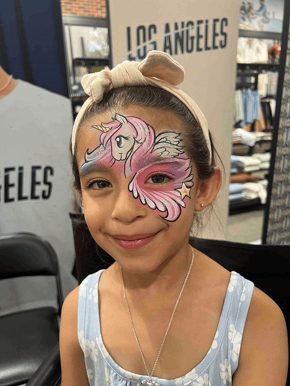 A young girl with a headband and blue dress has a colorful unicorn face painting covering one side of her face, standing near a balloon artist in Los Angeles twisting vibrant balloons.