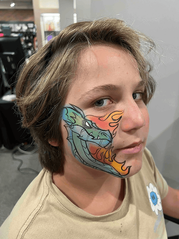 A person with light brown hair has a colorful dragon face painting covering their left cheek, featuring green, blue, and orange flames—perfect for making a statement at any corporate event alongside vibrant balloon installations.