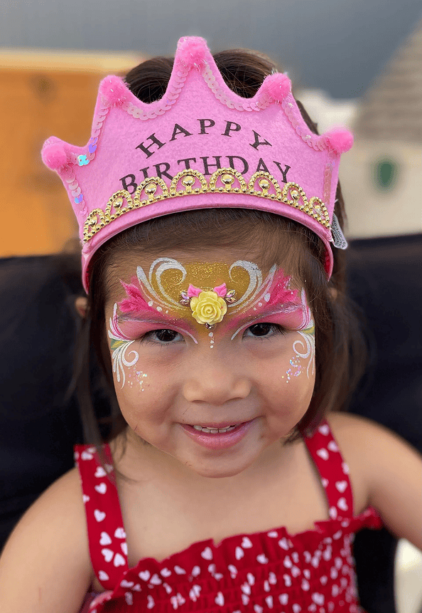 Young child wearing a pink "Happy Birthday" crown and red dress with heart patterns, smiling with elaborate pink and gold face paint, surrounded by joyful balloon decor.