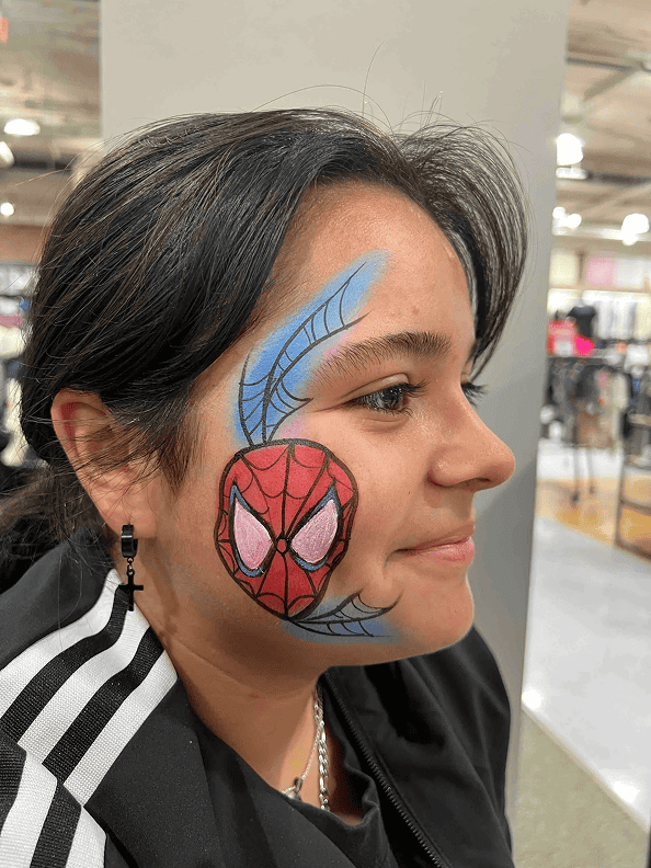 A person with dark hair and a black jacket has a painted Spiderman mask and blue web design on their cheek. They are indoors, possibly in a store decorated with colorful balloon installations.