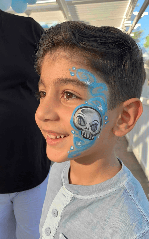 Smiling child with short brown hair, wearing a light blue shirt, has a blue and white skull face paint design on his cheek, enjoying a corporate event with colorful balloon decor.