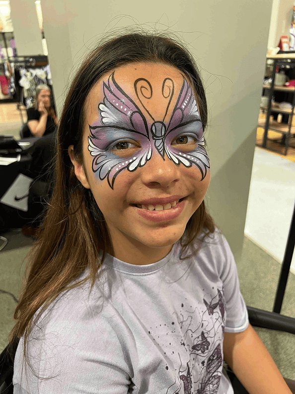 A girl with long hair smiles while wearing a purple and white butterfly face paint. She is sitting indoors, surrounded by colorful balloon decor, possibly in a Los Angeles store or event space.