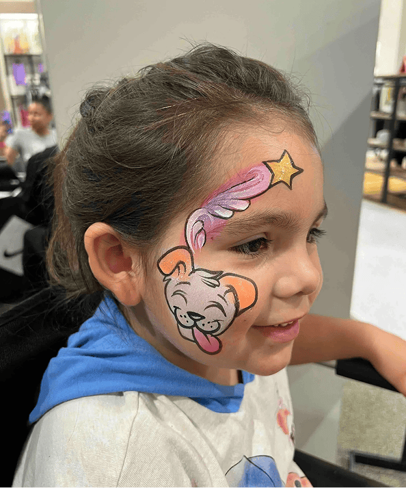 Young girl with face paint featuring a cartoon dog on her cheek and a pink shooting star with a gold star on her forehead, sitting indoors at a Los Angeles corporate event with colorful balloon decor.