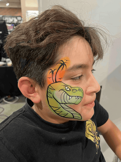 A child with brown hair has a green snake with fangs and an orange background painted on the side of their face by a talented balloon artist in Los Angeles.