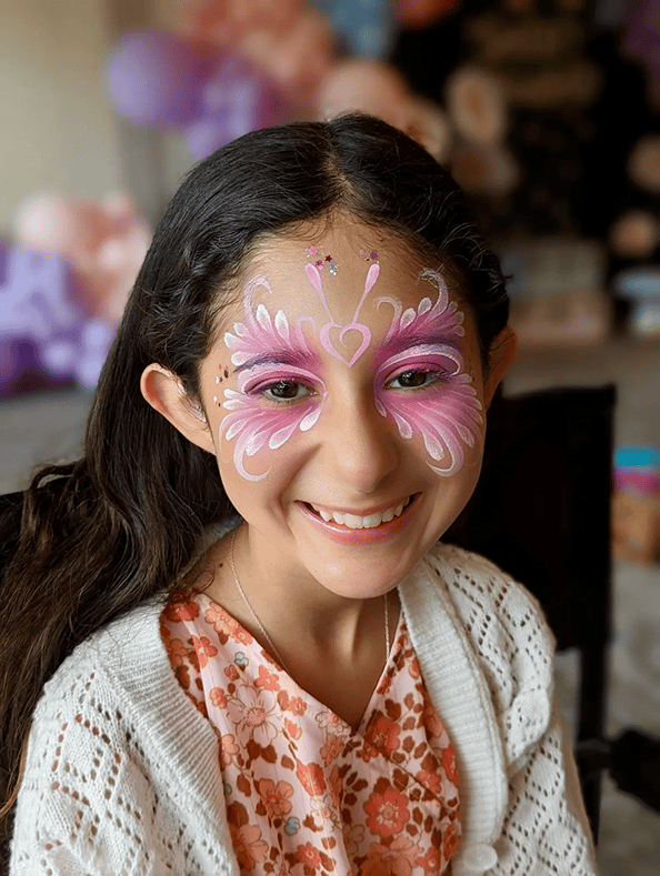 A girl with long dark hair smiles while wearing a white cardigan and floral dress. Her face is painted with pink and white butterfly designs, showcasing the work of a talented balloon artist at a Los Angeles event. Balloons blur in the background.