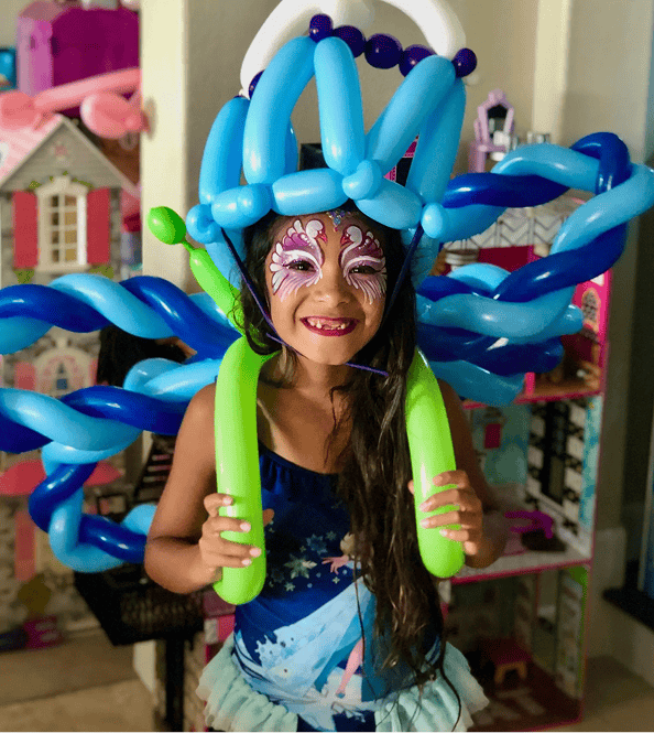 A smiling child with face paint and a blue balloon decor costume stands indoors in front of a dollhouse, ready to bring joy to any Los Angeles corporate event.