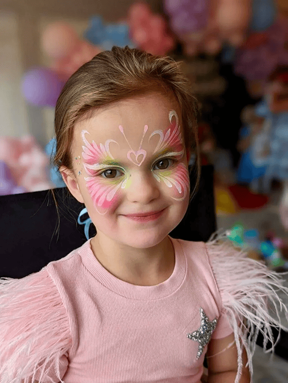 A young girl with brown hair wears a pink shirt with feathered sleeves and has colorful butterfly face paint. She is smiling and sitting indoors, surrounded by vibrant balloon decor.