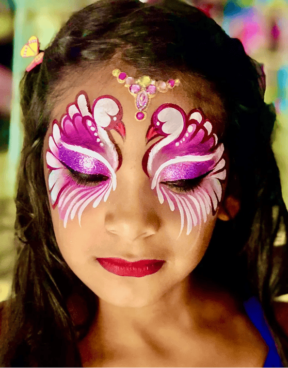 A child with long dark hair has her face painted with two pink and white swans forming a heart shape around her eyes, with gem accents on her forehead, at a Los Angeles event featuring a talented balloon artist.