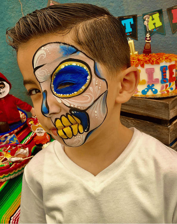 A young boy with neatly combed hair wears a white shirt and has his face painted as a colorful sugar skull at a decorated party table adorned with vibrant balloons.