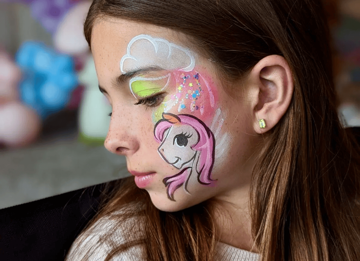 A girl with brown hair has her face painted with a unicorn, cloud, and colorful stars on her cheek and forehead at a Los Angeles event featuring stunning balloon decor.
