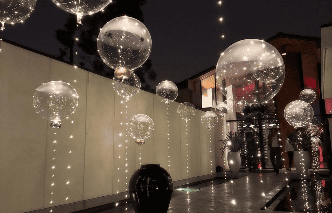 Large transparent balloons filled with string lights are suspended above a modern Los Angeles outdoor walkway at night, alongside a reflective water feature and potted plants, creating stunning balloon decor.