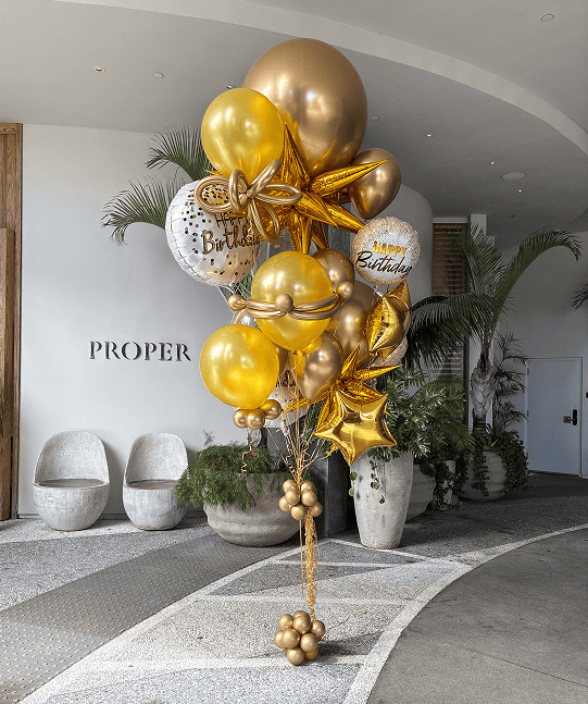 Gold and white birthday balloons, including star shapes and "Happy Birthday" messages, form a stylish balloon installation in a modern lobby with potted plants and curved seating—perfect for a festive corporate event atmosphere.