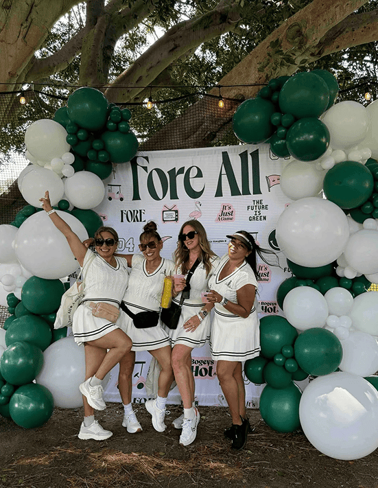 Four women in tennis outfits pose in front of a “Fore All” backdrop featuring striking balloon installations, outdoors at a corporate event in Los Angeles.