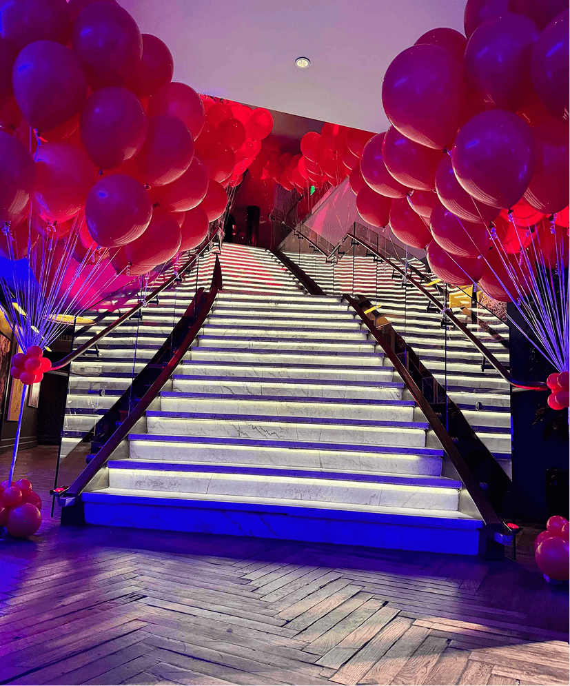 A grand staircase with illuminated steps, lined with bunches of red balloons on both sides, under vibrant pink and purple lighting—an impressive entrance perfect for a corporate event.