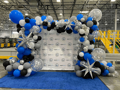 A striking balloon arch in blue, black, white, and silver with star decorations frames a step-and-repeat banner on a blue carpet—perfect for a stylish corporate event in Los Angeles’s industrial warehouse setting.