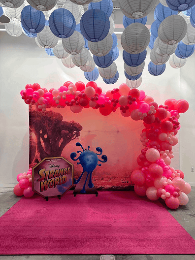 A "Disney Strange World" photo booth display in Los Angeles with pink and red balloons crafted by a balloon artist, a character cutout, and blue and white paper lanterns hanging from the ceiling.