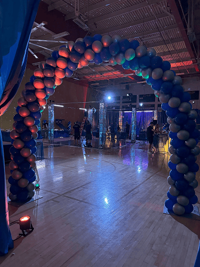 An arch made of blue and silver balloons, crafted by a skilled balloon artist, stands in a gymnasium decorated with lights and shiny streamers for an event in Los Angeles.