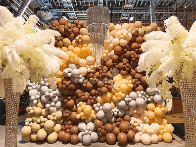 A decorative balloon wall in shades of gold, brown, and white stands behind crystal columns topped with white feather arrangements and orchids, showcasing elegant balloon decor indoors under exposed ceiling beams.
