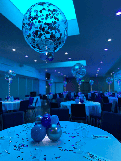 A banquet hall with round tables decorated with white cloths, elegant balloon decor in blue and silver, and confetti, all under soft blue lighting.