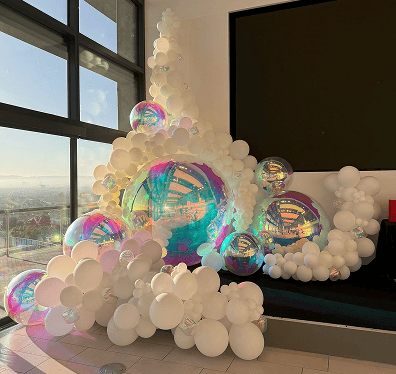 A cluster of large iridescent and white balloons creates elegant balloon decor near a window and a black bench in a modern Los Angeles room, perfect for a stylish corporate event.