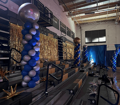 Decorated gym bleachers with blue and silver balloon installations, gold streamers, and metallic star-shaped decorations, likely set up for an event.