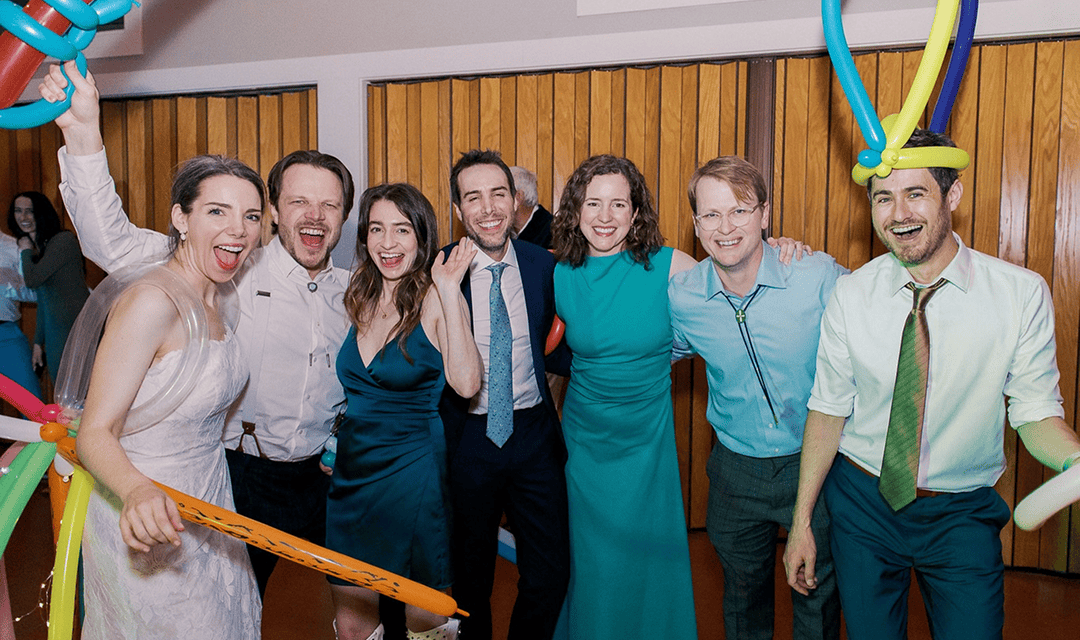 A group of seven adults in formal attire pose together indoors at a corporate event, some wearing balloon hats and holding balloons, smiling at the camera—likely creations from a skilled balloon artist.