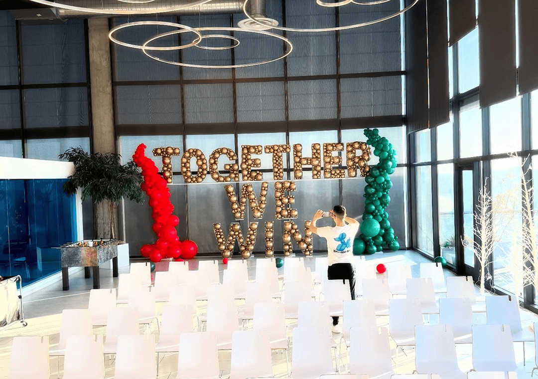 A person snaps a photo of a stage with “TOGETHER WE WIN” in lit marquee letters, vibrant balloon installations by a local Los Angeles balloon artist, and rows of white chairs in a bright, modern room.
