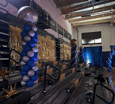 Rows of gym bleachers decorated by a balloon artist with blue and silver balloon columns, gold streamers, and metallic star-shaped ornaments for a festive corporate event.