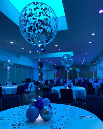 A decorated Los Angeles event hall with round tables, black chairs, and large confetti-filled balloon decor as centerpieces, all glowing under blue-tinted lighting.
