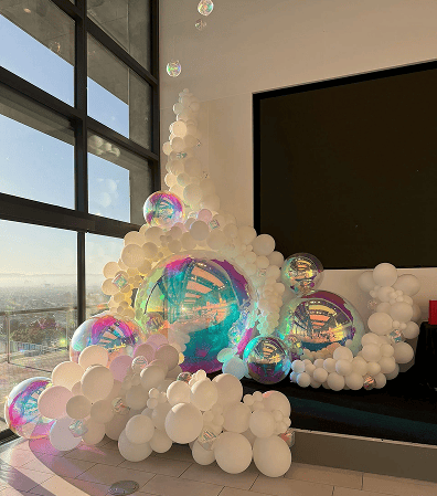 Large white and iridescent balloons are arranged in a decorative cluster near a corner window and a black display, showcasing the creativity of a skilled balloon artist in this modern Los Angeles indoor space.