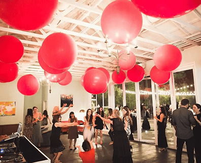 People dance in a well-lit room decorated with stunning balloon installations, including large red balloons hanging from the ceiling. The festive scene is perfect for a corporate event, with guests dressed in semi-formal attire.