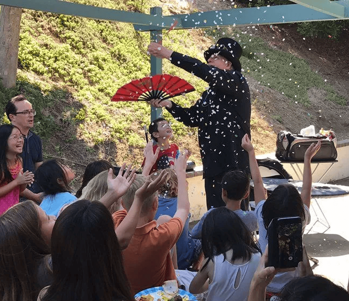A magician in Los Angeles entertains a group of children outdoors, holding a fan and creating a confetti effect as the audience watches and reaches up, surrounded by colorful balloon decor.