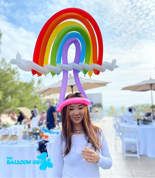 A woman stands outdoors at a party, wearing a colorful balloon hat shaped like a rainbow with clouds—crafted by a talented balloon artist—holding a drink, with decorated tables and umbrellas in the background.