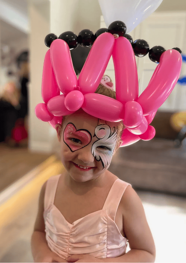 A young child wears a pink balloon crown created by a balloon artist and has face paint featuring a heart and swan design, dressed in a light pink outfit indoors.