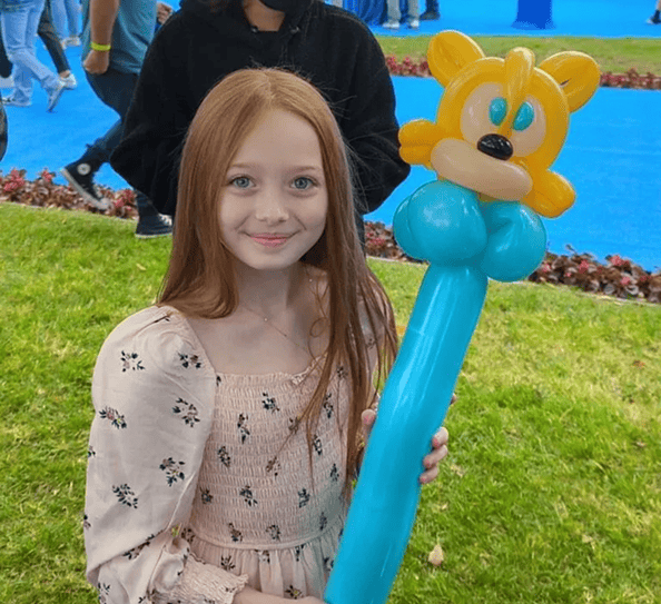 A girl with long red hair in a light dress holds a blue balloon animal shaped like a cartoon character while standing on grass at a corporate event featuring creative balloon installations in Los Angeles.