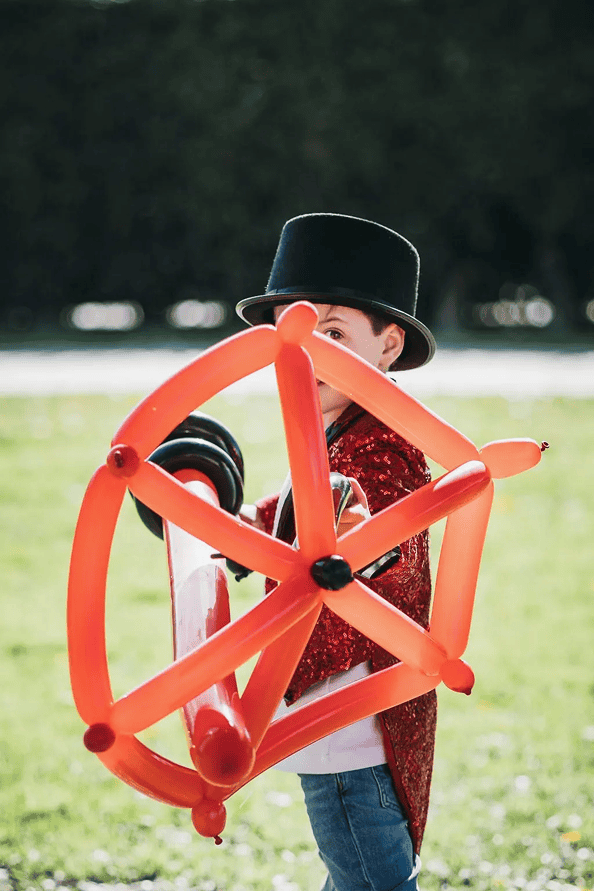 A child wearing a black top hat and red sequin cape holds colorful balloons shaped like a sword and star shield, standing outdoors on grass in Los Angeles.