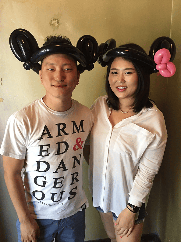 Two people stand side by side, both wearing black balloon hats shaped like mouse ears—creations by a talented Los Angeles balloon artist. One hat is topped with a pink bow. They are indoors, posed against a plain wall.