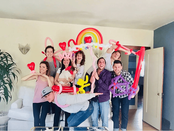 A group of eight people pose indoors with colorful balloon decor, including hearts, animals, and a rainbow, smiling and making playful gestures as they enjoy the creative balloon installations.