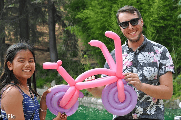 Two people stand outdoors, smiling and holding a pink balloon sculpture shaped like a bicycle, showcasing the creativity of a balloon artist. Trees and greenery fill the background.