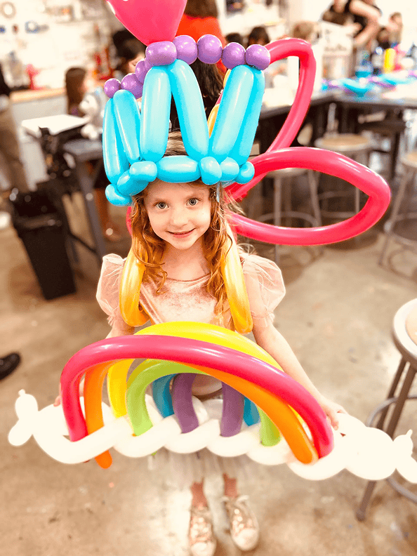 A young girl wearing a pink dress and a colorful balloon costume, created by a talented balloon artist, stands indoors on a concrete floor in Los Angeles.